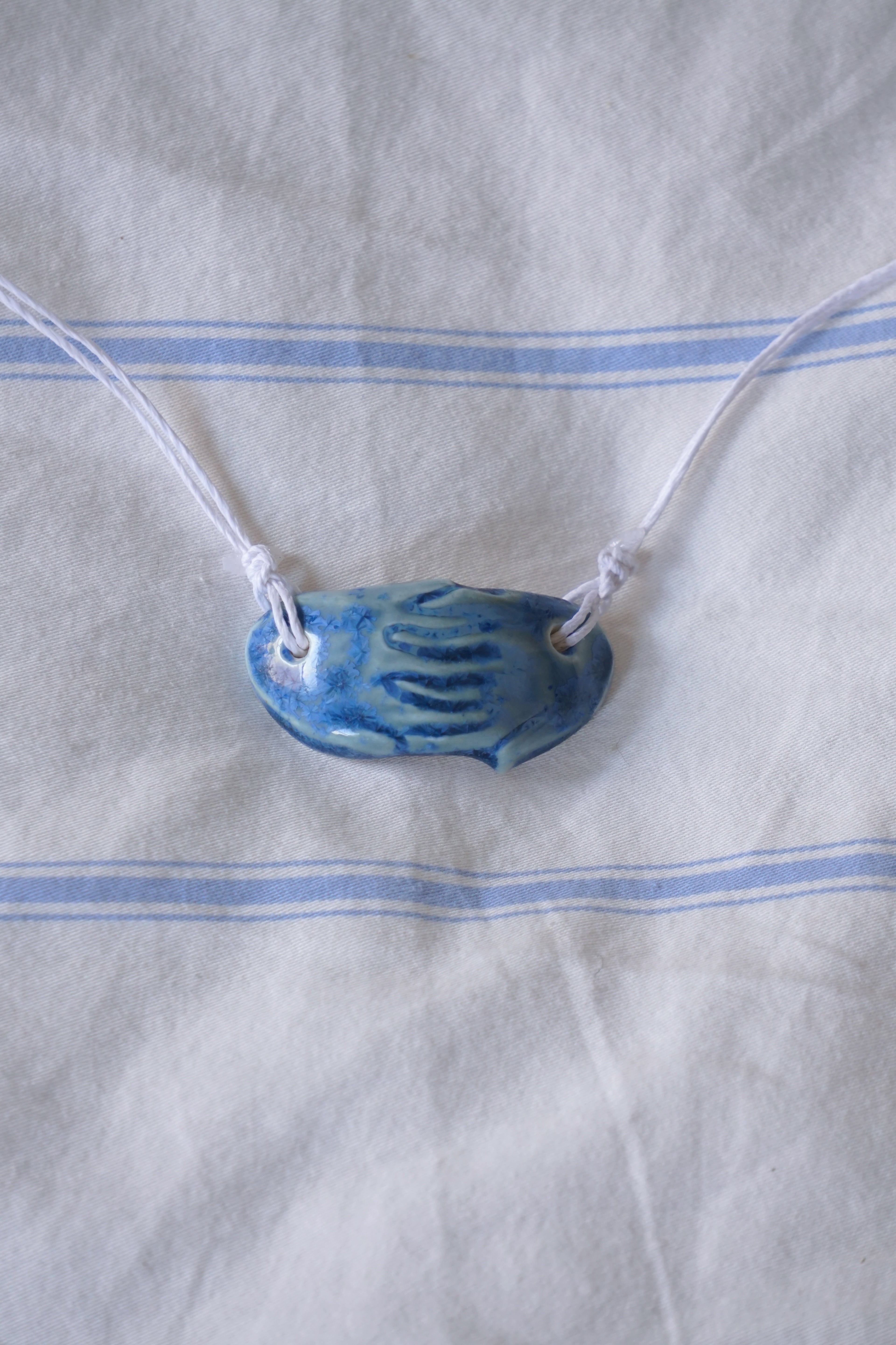 Blue ceramic pendant on a white cord against a white fabric background with blue stripes